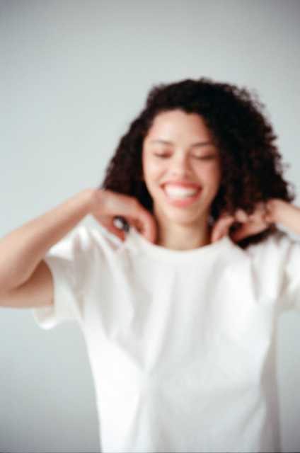 Woman with curly hair smiling and touching the collar of her crew neck tee against a light gray background
